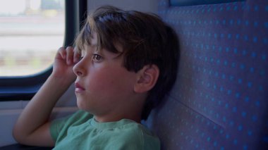 Çocuk sessizce tren penceresinden dışarı bakar, el şakağına, vücudunu koltuğa sıkıştırır, ciddi ve odaklanmış bir şekilde, manzara dışarı doğru koşarken.