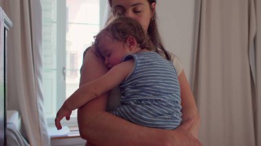 Anne pencerenin önünde duruyor uyuyan bebeği göğsüne yaslıyor, doğal ışık perdeleri ve camdan görülebilen bir bina.