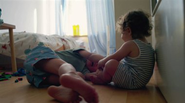 Yürüyen çocuk, büyük kardeşinin yatıp, yatak odasında oynamasını seyrediyor. Camdan gün ışığı akıyor. Etrafa saçılan renkli oyuncaklar.