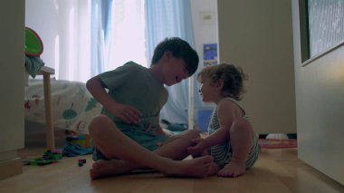 İki küçük kardeş, tahta zeminde güneşli yatak odasında birlikte oynuyorlar. Bebek, oyuncaklarla ve gün ışığıyla çevrili büyük çocuğu sıcak bir şekilde kucaklıyor.