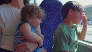 Bebek annesinin kollarında dik oturup otobüse bakarken, büyük çocuk pencereye yaslanıyor ve geçen şehir manzarasını seyrediyor.