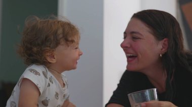 Anne ve bebek, yeni yürümeye başlayan çocuk su içtikten sonra gülüyorlar. Neşeli bir bağ ve neşe anı. Birbirlerine gülerek bakıyorlar.