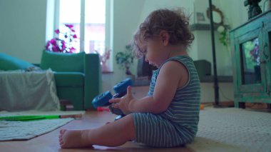 Küçük çocuk içeride oturmuş, mavi oyuncak robotu çeviriyor. Odaklanıyor ve merak ediyor. Etrafı rahat mobilyalarla çevrili ve pencereden içeri ışık giriyor.