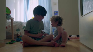 İki küçük kardeş, tahta zeminde güneşli yatak odasında birlikte oynuyorlar. Bebek, oyuncaklarla ve gün ışığıyla çevrili büyük çocuğu sıcak bir şekilde kucaklıyor.