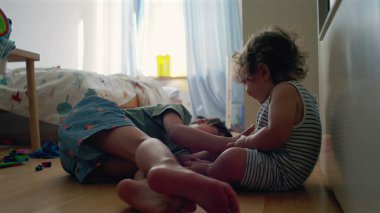 Yürüyen çocuk, büyük kardeşinin yatıp, yatak odasında oynamasını seyrediyor. Camdan gün ışığı akıyor. Etrafa saçılan renkli oyuncaklar.