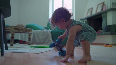 İçeride ahşap zemine çömelmiş küçük çocuk, yoğun merak ve denge ile mavi oyuncak robotu dikkatlice inceliyor, rahat oturma odası mobilyaları ile çevrili