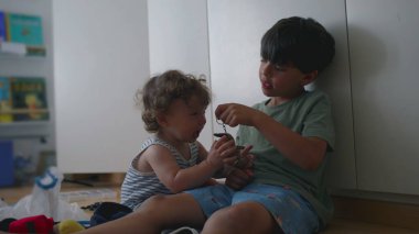 Büyük kardeş, bebek kardeşlerin kafasının yanında anahtarlık oyuncağını sallarken yeni yürümeye başlayan ördekler ve kalkanlar arka planda kitaplarla birlikte çocuk odasında yerde oturuyorlar.