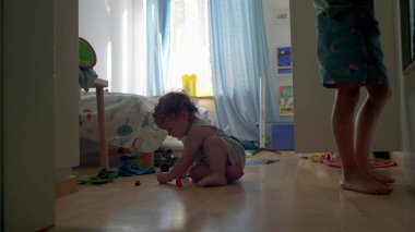 Yürüyen çocuk yatak odasında tek başına oynuyor. Oyuncak parçaları tam konsantrasyon, parlak pencere ışığı ve arka plandaki eğlenceli oda detaylarıyla inceliyor.