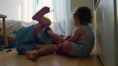 Yürüyen çocuk, büyük kardeşinin yatıp, yatak odasında oynamasını seyrediyor. Camdan gün ışığı akıyor. Etrafa saçılan renkli oyuncaklar.