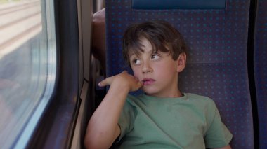 Trenin penceresinde oturan çocuk, el ele tutuşmuş, yolculuk sırasında düşünceli bir ifadeyle dışarı bakıyor, samimi çocukluk anı yansıması ve durgunluk.