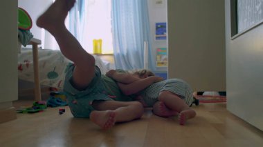 İki küçük kardeş, tahta zeminde güneşli yatak odasında birlikte oynuyorlar. Bebek, oyuncaklarla ve gün ışığıyla çevrili büyük çocuğu sıcak bir şekilde kucaklıyor.