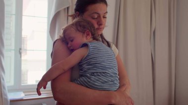 Anne pencerenin önünde duruyor uyuyan bebeği göğsüne yaslıyor, doğal ışık perdeleri ve camdan görülebilen bir bina.