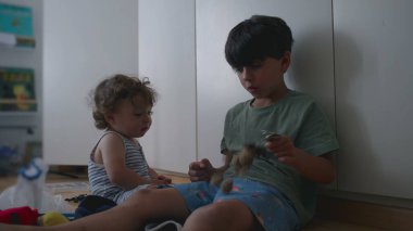 Büyük kardeş anahtarlık pelüş oyuncakla oynuyor. Küçük kardeş merakla izliyor. Her ikisi de arka planda kitap ve oyuncaklarla dolu, rahat bir odada, ahşap zeminde oturuyor.