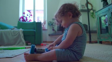 Küçük çocuk içeride oturmuş, mavi oyuncak robotu çeviriyor. Odaklanıyor ve merak ediyor. Etrafı rahat mobilyalarla çevrili ve pencereden içeri ışık giriyor.