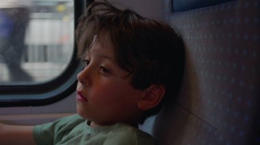 Çocuk sessizce tren penceresinden dışarı bakar, el şakağına, vücudunu koltuğa sıkıştırır, ciddi ve odaklanmış bir şekilde, manzara dışarı doğru koşarken.