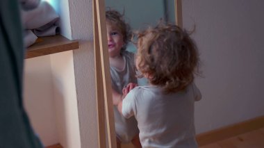 Küçük çocuk aynaya sarılıp kameraya doğru yürümeden önce arkasını dönüyor, kapalı alanda hareket, denge ve erken keşifler gösteriyor.