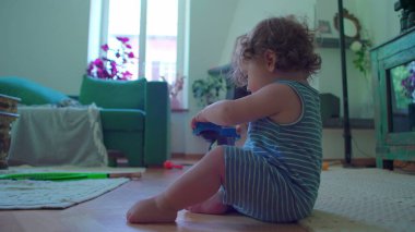 Küçük çocuk içeride oturmuş, mavi oyuncak robotu çeviriyor. Odaklanıyor ve merak ediyor. Etrafı rahat mobilyalarla çevrili ve pencereden içeri ışık giriyor.