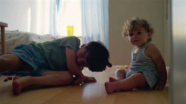 Bebek ve büyük kardeş yatak odasında dinleniyor, gülümsüyor ve doğal ışık ve dağınık oyuncaklar arasında oynuyor, evde sessiz bir anda bağ kuruyorlar.