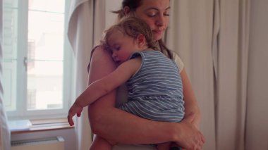 Anne pencerenin önünde duruyor uyuyan bebeği göğsüne yaslıyor, doğal ışık perdeleri ve camdan görülebilen bir bina.