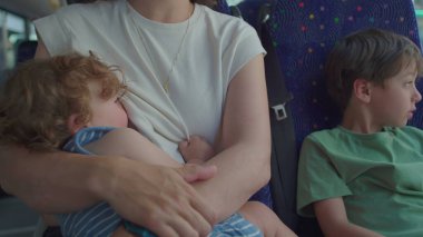 Anne otobüste bebeği emzirirken, büyük kardeş onların yanında el kol hareketleri yapıyor, şehir içi seyahatler sırasında şablonlu koltukta oturuyor.