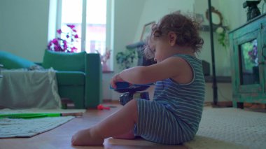 Küçük çocuk içeride oturmuş, mavi oyuncak robotu çeviriyor. Odaklanıyor ve merak ediyor. Etrafı rahat mobilyalarla çevrili ve pencereden içeri ışık giriyor.