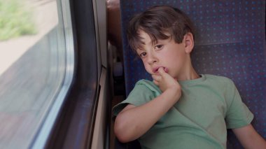 Trenin penceresinde oturan çocuk, el ele tutuşmuş, yolculuk sırasında düşünceli bir ifadeyle dışarı bakıyor, samimi çocukluk anı yansıması ve durgunluk.