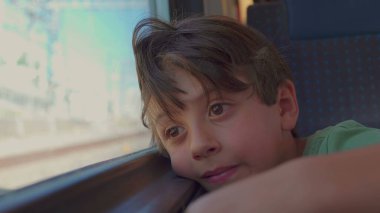 Genç çocuk trenin penceresinde dinleniyor, hüzünlü bir ifadeyle dışarı bakıyor, güneş ışığı yüzünü ve saçlarını aydınlatıyor gezinti sırasında manzara bulanıklaşırken