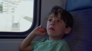 Dalgın çocuk mavi tren koltuğuna uzanıyor, sessizce pencereden dışarı bakıyor, güneş ışığı altındaki manzara dışarı doğru sürüklenirken, el şakağına hafifçe dokunuyor.