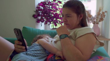 Anne akıllı telefona bakarken bebeği emziriyor, pencerenin yanındaki koltukta oturuyor ve arka planda doğal ışık ve bitkiler var.