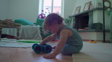 İçeride ahşap zemine çömelmiş küçük çocuk, yoğun merak ve denge ile mavi oyuncak robotu dikkatlice inceliyor, rahat oturma odası mobilyaları ile çevrili