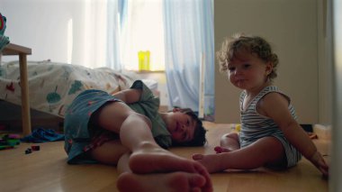 Bebek ve büyük kardeş yatak odasında dinleniyor, gülümsüyor ve doğal ışık ve dağınık oyuncaklar arasında oynuyor, evde sessiz bir anda bağ kuruyorlar.
