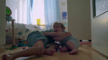 İki küçük kardeş, tahta zeminde güneşli yatak odasında birlikte oynuyorlar. Bebek, oyuncaklarla ve gün ışığıyla çevrili büyük çocuğu sıcak bir şekilde kucaklıyor.