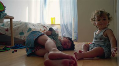 Bebek ve büyük kardeş yatak odasında dinleniyor, gülümsüyor ve doğal ışık ve dağınık oyuncaklar arasında oynuyor, evde sessiz bir anda bağ kuruyorlar.