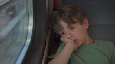 Düşünceli çocuk tren koltuğunda tek başına oturur, kafası dinlenir, yorgun bir ifadeyle pencereden dışarı bakar, dışarıda hareketli izler bulanıklaşır.