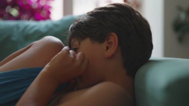 Kanepede kıvrılırken kumaşı ısıran çocuğun yakın çekimi, iç gözlem havası ve ortam aydınlatması evdeki sessiz yansımanın atmosferini artırıyor.