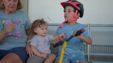 Yürüyen çocuk, aktiviteden ya da sürmekten bir an önce yakınlarda büyükannesiyle birlikte kaskını takıp scooter tutan büyük çocuğun yanında oturur.