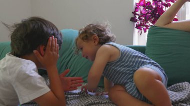 Kıvırcık saçlı çocuk, ablasının yanında şakacı bir şekilde yuvarlanırken anne arka planda dinleniyor, parlak oturma odası kuruluyor.