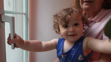 Büyükannesinin tuttuğu kıvırcık saçlı çocuk, dışarıyı merakla izlerken kapı koluna uzanıyor, evin içini hafifçe aydınlatıyor.