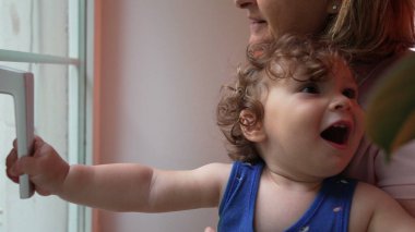 Büyükannesinin tuttuğu kıvırcık saçlı çocuk, dışarıyı merakla izlerken kapı koluna uzanıyor, evin içini hafifçe aydınlatıyor.