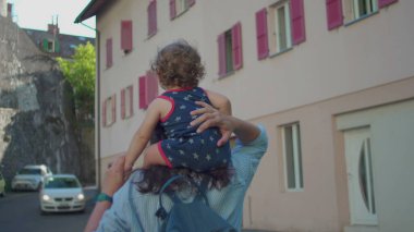 Annem, güneşli bir öğleden sonra pastel binalar ve kırmızı pencere panjurlarıyla dar Avrupa caddelerinde yürürken omuzlarında bebek taşır.