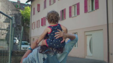 Annem, güneşli bir öğleden sonra Pastel binalar ve pencerelerde kırmızı panjurlarla kaplı sakin bir sokakta yürürken omuzlarında bebek taşır.