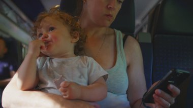Annem trende kıvırcık saçlı bir çocuğu kucağında tutuyor, bebek atıştırmalıkları. Kadın telefonlarını kontrol ediyor, arka planda oturan yolcuları görüyor.
