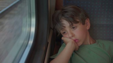 Düşünceli çocuk tren koltuğunda tek başına oturur, kafası dinlenir, yorgun bir ifadeyle pencereden dışarı bakar, dışarıda hareketli izler bulanıklaşır.