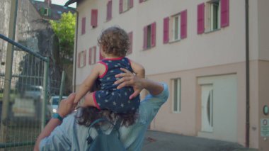 Annem, güneşli bir öğleden sonra Pastel binalar ve pencerelerde kırmızı panjurlarla kaplı sakin bir sokakta yürürken omuzlarında bebek taşır.