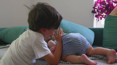 Kıvırcık saçlı çocuk, ablasının yanında şakacı bir şekilde yuvarlanırken anne arka planda dinleniyor, parlak oturma odası kuruluyor.