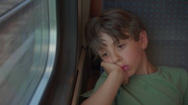 Düşünceli çocuk tren koltuğunda tek başına oturur, kafası dinlenir, yorgun bir ifadeyle pencereden dışarı bakar, dışarıda hareketli izler bulanıklaşır.