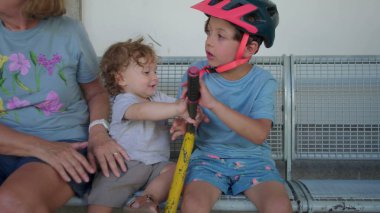 Yürüyen çocuk, aktiviteden ya da sürmekten bir an önce yakınlarda büyükannesiyle birlikte kaskını takıp scooter tutan büyük çocuğun yanında oturur.