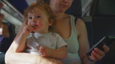 Annem trende kıvırcık saçlı bir çocuğu kucağında tutuyor, bebek atıştırmalıkları. Kadın telefonlarını kontrol ediyor, arka planda oturan yolcuları görüyor.