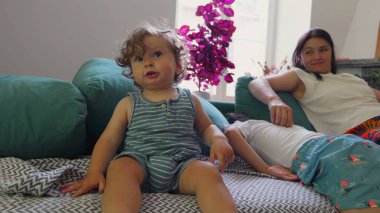 Küçük çocuk koltukta dik otururken anne uzanır ve ağabeyi yastıklarda uzanır. Huzurlu, rahat bir oturma odasında huzurlu bir aile anı.