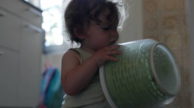 Bebek, mutfak zemininde oturmuş ev eşyalarıyla uğraşırken salata fırıldağı kapağında el sıkışıyor.